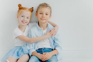 Friendly small girl and boy hug and sit on floor against white background, have positive expressions, feel satisfied after playing together, copy space. Ginger female kid embraces brother with love photo