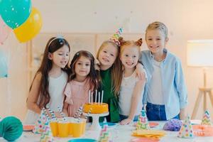 Horizontal shot of group of little children gather together to celebrate birthday, embrace and pose at camera, prepare for special occasion, stand near table with cake, paper cups, party caps photo