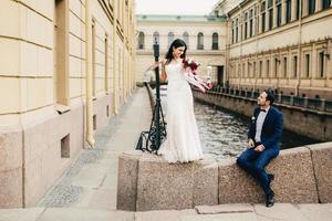 Handsome bridegroom sits on bridge looks at his beautiful bride who stands up, says compliments to her, enjoy togetherness, have unforgettable moments together, Romantic people and wedding concept photo