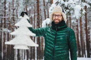 Pleasant looking bearded male dressed in warm clothes, holds artificial fir tree, stands against beautiful trees covered with snow, has cheerful positive expression, breathes fresh winter air photo