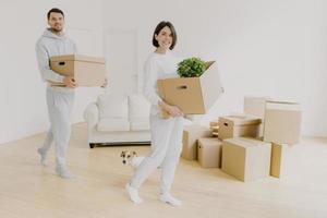 Horizontal shot of happy young couple move in new home, buy real estate, busy with carrying personal belongings in carton boxes, pose in empty room, dog is near on floor. New beginning concept photo