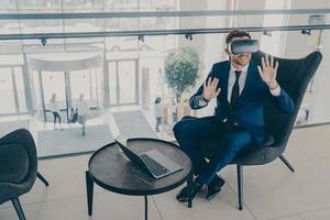 Smiling office worker sitting in company center lobby with laptop and VR headset goggles photo