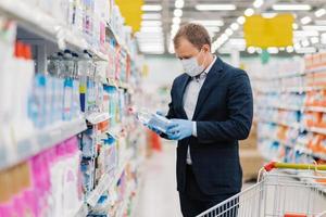 Shot of adult man chooses detergent in housekeeping store, reads label and instruction how to use product, wears medical mask and gloves during coronavirus pandemic, avoids risk to catch virus photo