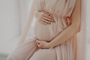 Cropped image of faceless woman touches her abdomen anticipates for baby wears festive dress poses indoor. Women and pregnancy concept photo