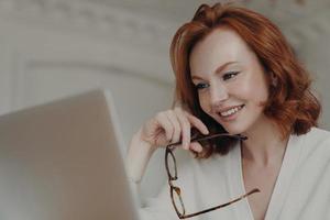 Professional smiling ginger female web designer uses application on laptop computer for creating project work, makes online research and browses internet, holds spectacles, has happy expression photo