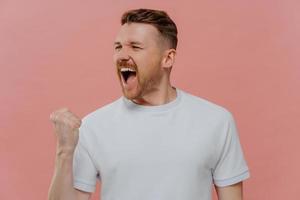 el hombre exitoso emocional grita de alegría aprieta el puño con el triunfo hace que el gesto de la bomba celebre el triunfo vestido con una camiseta blanca casual aislada sobre fondo rosa. concepto de lenguaje corporal foto