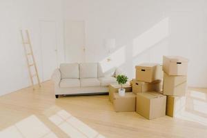 Spacious room with sofa, piles of cardboard boxes and ladder, white walls, with no people, personal belongings, domestic flower in pot, floor lamp. Loan mortgage and delivery service concept photo