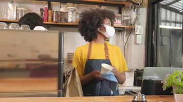 la barista afroamericana trabaja limpiando la taza de café, mirando a través de la ventana del café, esperando a los clientes en un nuevo servicio de estilo de vida normal, el impacto comercial de la cuarentena pandémica covid-19. video