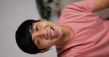 Vertical POV Video Call Portrait of Asian young man talking to friends or colleagues while standing inside in living room. Smiling man waving hand in home.