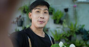 porträtt av en glad ung asiatisk manlig trädgårdsmästare som säljer online på sociala medier och tittar på kameran i trädgården. man selfie med mobiltelefon. hem grönska, försäljning online och hobby. video