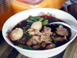 fideos tailandeses, fideos pequeños con cerdo estofado, albóndigas, hígado de cerdo y sopa de sangre en un tazón blanco, palillos de madera y cuchara, sazone la comida con el condimento, comida tailandesa deliciosa foto