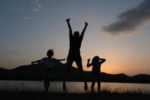 Silhouette of little daughters and mother jumping by the lake at sunset. Happy family playing together in the garden. Healthy summer activity for children. Funny time with family.