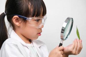 Portrait of a little girl in glasses holding a magnifying glass looking at leaves in researcher or science uniform on white background. Little scientist. photo