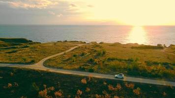 Above view of white car riding country road, turning to sea coast at warm sunset. Panoramic view of scenic valley with suv on empty road with marine sunset on background . Concept of destination video