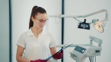 Caucasian female dentist doctor in dental office in protective transparent mask and folder in hands. Girl dentist looking at camera. video