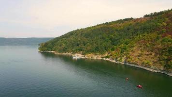 Above view of picturesque seashore and sea surface, with small ship mooring ashore at daytime. Calm sea washing green woodland hills against clear blue sky in warm summer day. Concept of landscape video