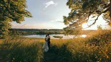 Side view of attractive bride and groom standing, hugging on field in setting sun rays. Camera approaching to wedding couple while they kissing with beautiful lake on background. Concept of wedding video