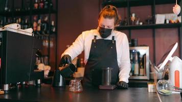 Female barista in protective mask preparing coffee, pouring hot drink into cup in cafe. Brunette girl with ponytail making pourover, holding coffee pot at bar. Concept of working place video