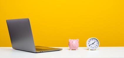 white desk with laptop clock and pink piggy bank yellow background with copy space photo