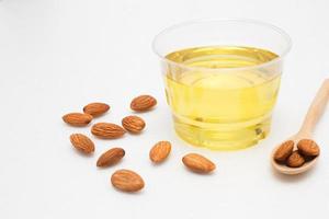 Yellow almond oil in a clear bowl with a wooden spoon next to it and several almonds on a white background. photo