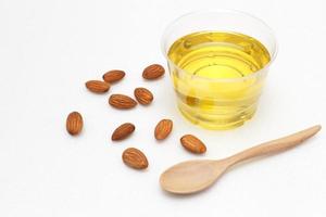 Yellow almond oil in a clear bowl with a wooden spoon next to it and several almonds on a white background. photo