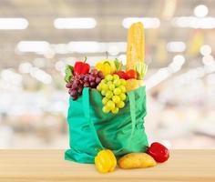 Fresh fruits and vegetables in reusable green shopping bag on wood table top with supermarket grocery store blurred defocused background with bokeh light photo