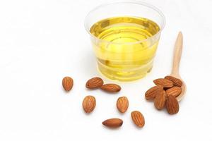 Yellow almond oil in a clear bowl with a wooden spoon next to it and several almonds on a white background. photo