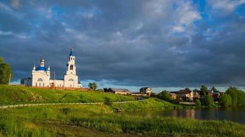 una antigua iglesia ortodoxa y un pueblo en la orilla del río. sol puesta de sol y nubes negras. foto