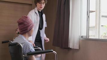 Uniformed young Caucasian female therapy doctor pushes wheelchair male patient to window, supports and motivates recovery from cancer illness after chemo medical treatment in hospital inpatient room. video