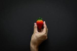 Hand holding strawberry on dark background for minimalist flat lay black food concept. photo