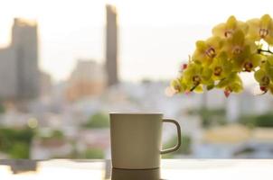 A cup of coffee on table with phalaenopsis orchid and city background in morning.