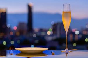 A glass with vapour of cold Champagne with dish for dining put on table with colorful city bokeh lights background.