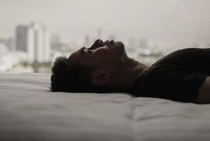 Asian man feels sad alone on bed with city background. Stay home, depression and loneliness concept. photo