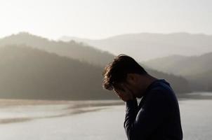 Asian miserable depressed man stand alone with mountain and lake background. Depression and mental health concept. photo