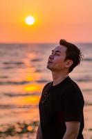 hombre asiático mirando al cielo con puesta de sol en el fondo de la playa. expresión positiva de la cara de la salud mental y la emoción. foto