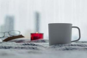 una taza de café se pone en la cama con una vela aromática y un libro en la temporada de invierno con un fondo borroso de la ciudad. foto