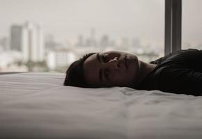 el hombre asiático se siente triste solo en la cama con el fondo de la ciudad. quedarse en casa, depresión y concepto de soledad. foto