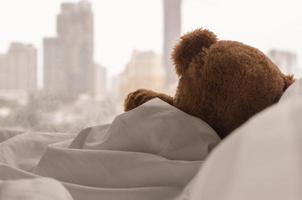 Teddy bear sleeping alone on bed with white pillow and blanket facing to window with rain drop in the lonely day. photo