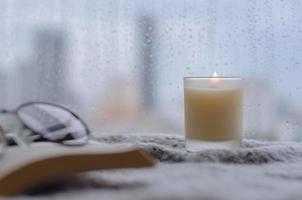 Burning aroma candle puts near by window that have rain drop in monsoon season with blurred city background. photo