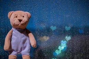 Teddy bear standing at window when raining with colorful love shape bokeh lights. photo