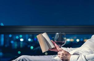 el hombre que se relaja en la habitación leyendo un libro con una copa de vino tinto y el fondo de las luces de la ciudad. foto