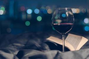 una copa de vino tinto puesta en la cama con un libro y un colorido bokeh del fondo de las luces de la ciudad. foto