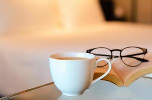 Selective focus of a white cup coffee puts on glass table with book and spectacles in bedroom. Relaxing at home concept. photo