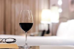 una copa de vino tinto con gafas y un libro sobre la mesa en el dormitorio. relajarse en el concepto de casa. foto