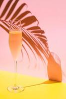 A glass of cold Champagne with vapour that has coconut leaf shadow from sun light on pink and yellow background. Summer concept. photo