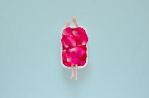 Doll bathing with rose petals in bathtub on pastel blue background. Top view minimal beauty summer concept. photo