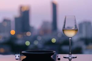 una copa de vino blanco con un plato para cenar puesto en la mesa con un colorido fondo de luces de la ciudad. foto