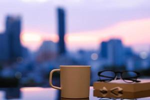 A cup of coffee with spectacles on book and ear pod put on table with city background and dawn sky. Stay home concept. photo