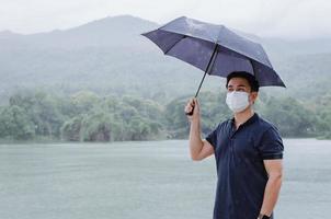 hombre asiático con mascarilla y paraguas para protegerse de enfermarse y mojarse cuando llueve en la temporada del monzón. foto