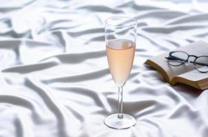 una copa de vino rosado sobre tela satinada ondulada con libro y gafas. quedarse en casa concepto. foto